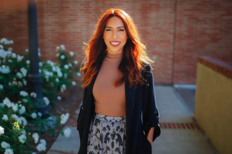 Drea Letamendi posing outdoors in natural light; she is smiling warmly, dressed in a neutral-toned top and black jacket, set against a backdrop of greenery and a brick wall.