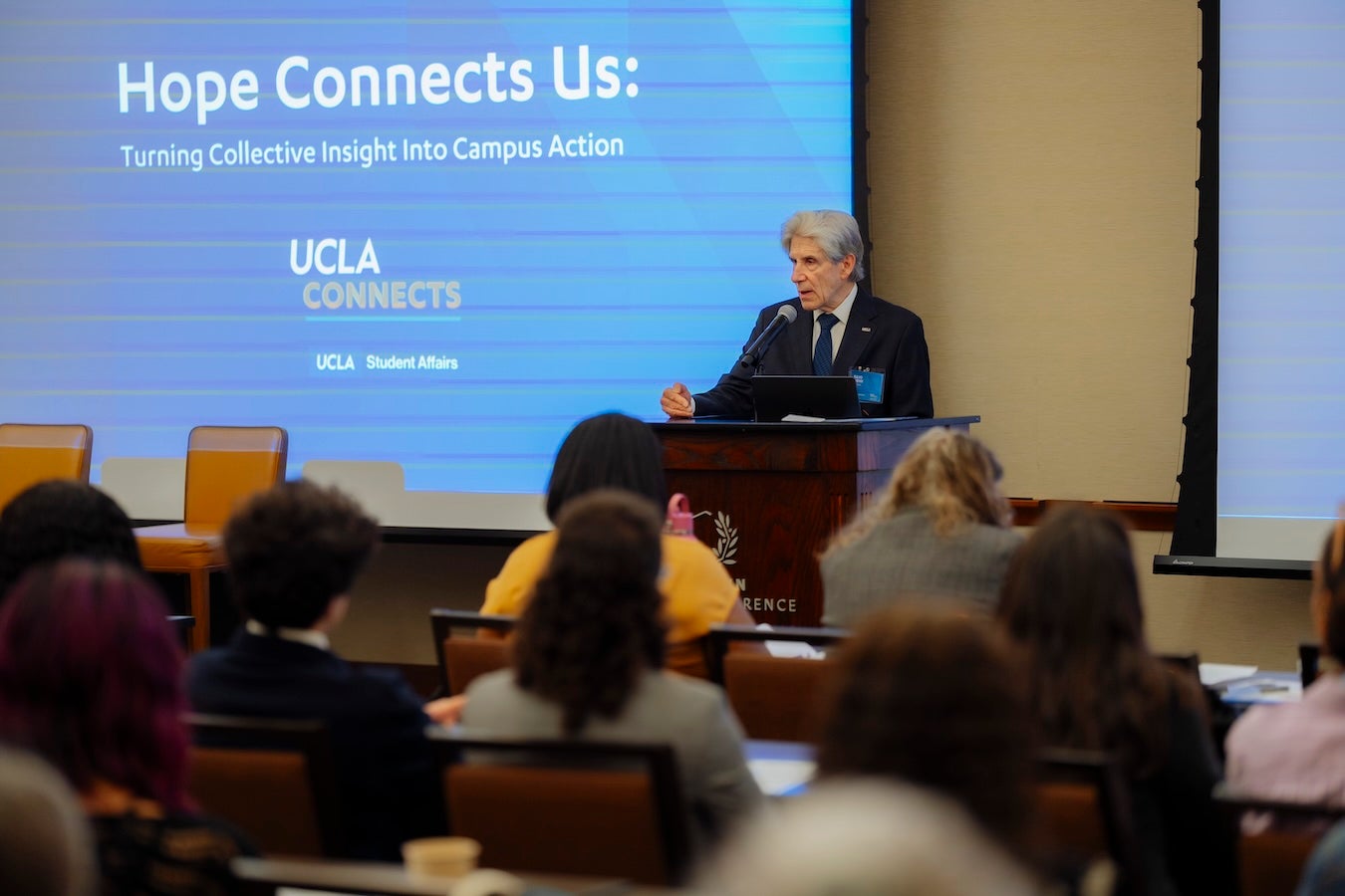 UCLA Chancellor Julio Frenk stands at a wooden podium speaking into a microphone before an audience of attendees seated at tables in a conference room. Behind him, a large blue projection screen displays the text Hope Connects Us.